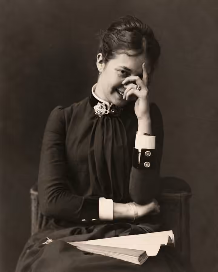 Original monochrome photograph of a seated smiling woman holding her hand over part of her face, with papers across her lap